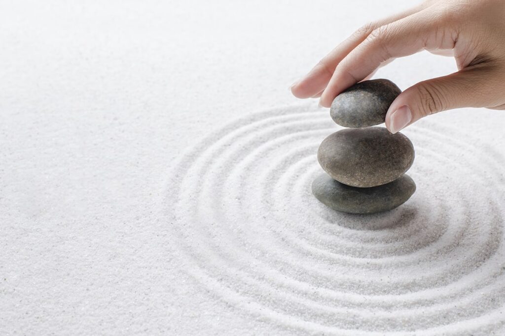 A person's hand carefully places the top stone on a stack of three smooth, dark gray zen stones (cairn), centered on white sand raked into concentric circles in a Japanese rock garden (Karesansui) style.